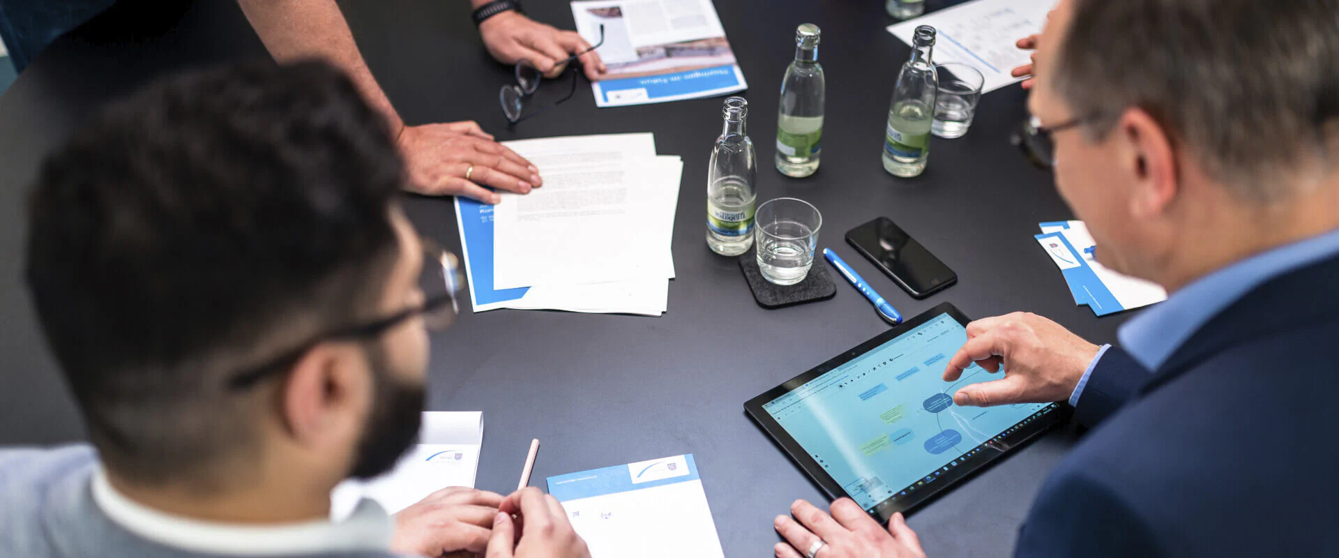 Weiterbildung Bildbeschreibung / description: Menschen beraten sich an einem Tisch mit einem Tablet und Unterlagen.